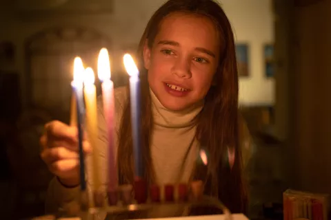 A smiling young girl lights colorful Hanukkah candles on a menorah in a dim, warmly lit room